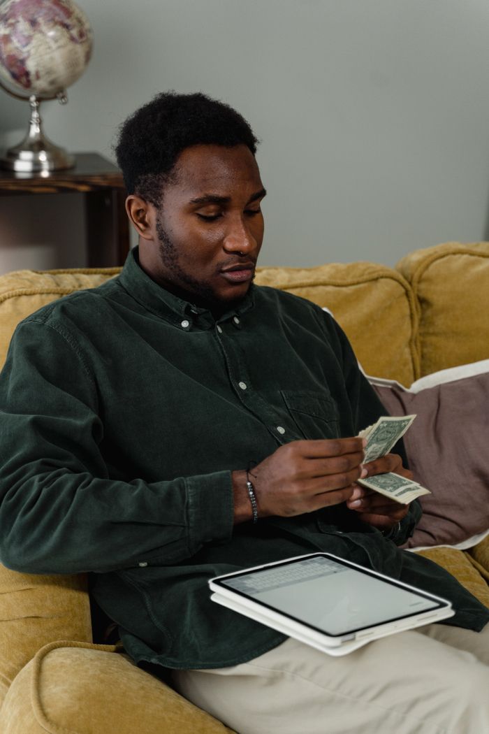 A businessman counting cash while seated on a sofa with a tablet at hand, reflecting on financial management.